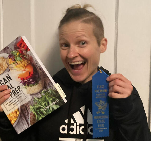 Major Award EG, the baker, with an excited smile holding up her prizes (a cookbook and blue ribbon) from winning the 2024 MN State Fair Vegan Main Dish Competition